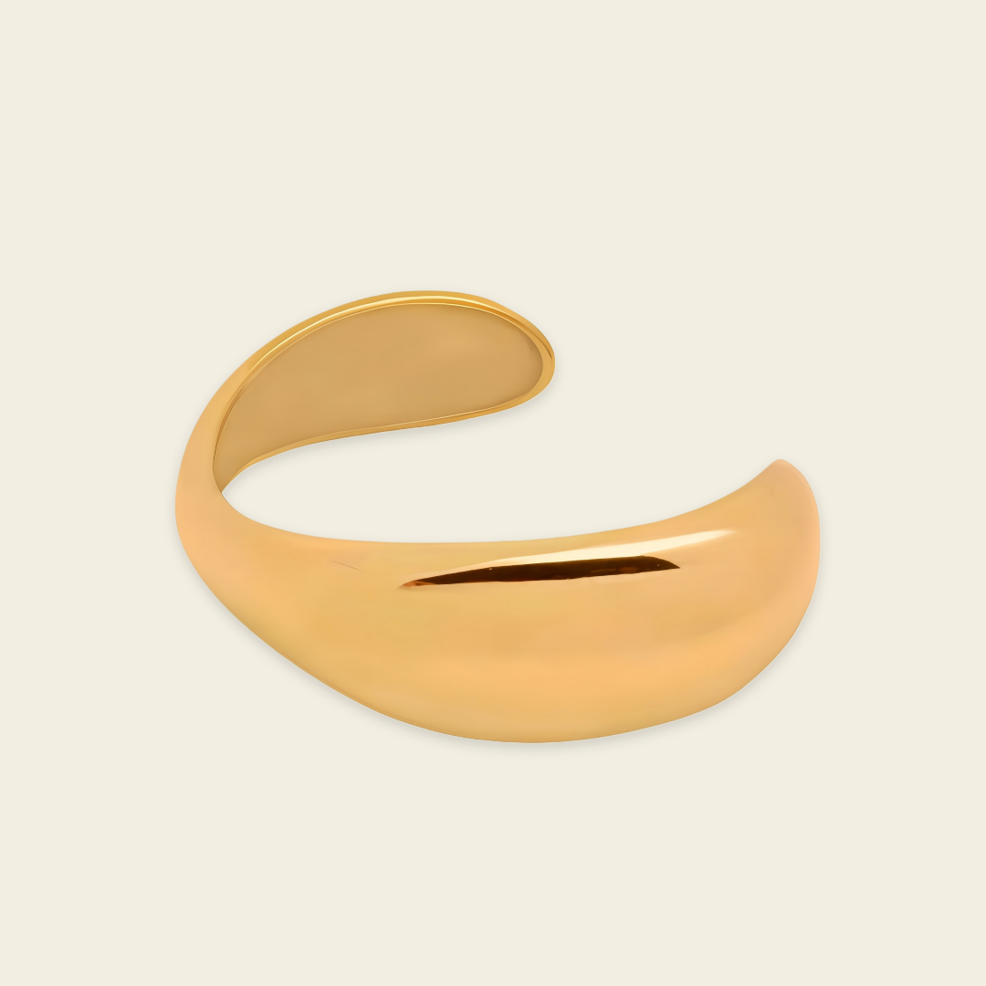 A polished gold-toned open cuff bangle with a smooth, curved organic shape, displayed on a light cream background.