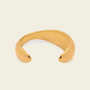 A polished gold-toned open cuff bangle with a smooth, organic curved shape, displayed on a solid light cream background.