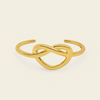 A polished gold open cuff bracelet featuring an integrated infinity knot design, presented on a clean, solid cream background.