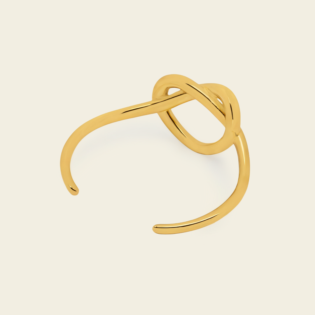 A gold-toned open cuff ring with a sculptural, intertwined knot-like design, shown on a solid light cream background.