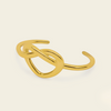 A polished gold open cuff ring with an abstract, interlocking knot-like design, shown on a solid light beige background.