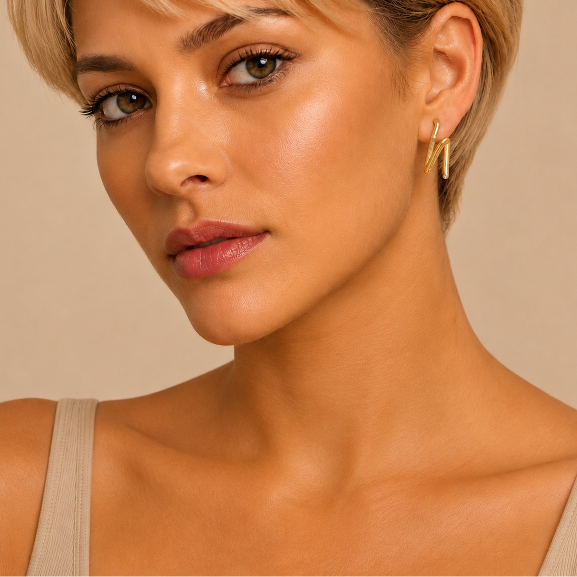 Close-up of a woman with short blonde hair wearing a pair of gold geometric hoop earrings against a warm beige background.