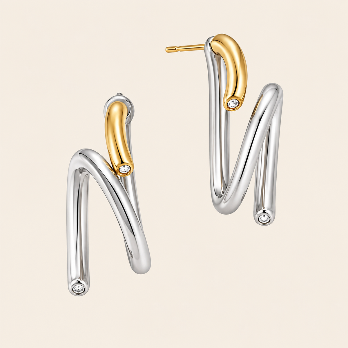 A pair of modern, abstract curved drop earrings in polished silver and yellow gold, each featuring a small clear crystal accent, shown on a light beige background.