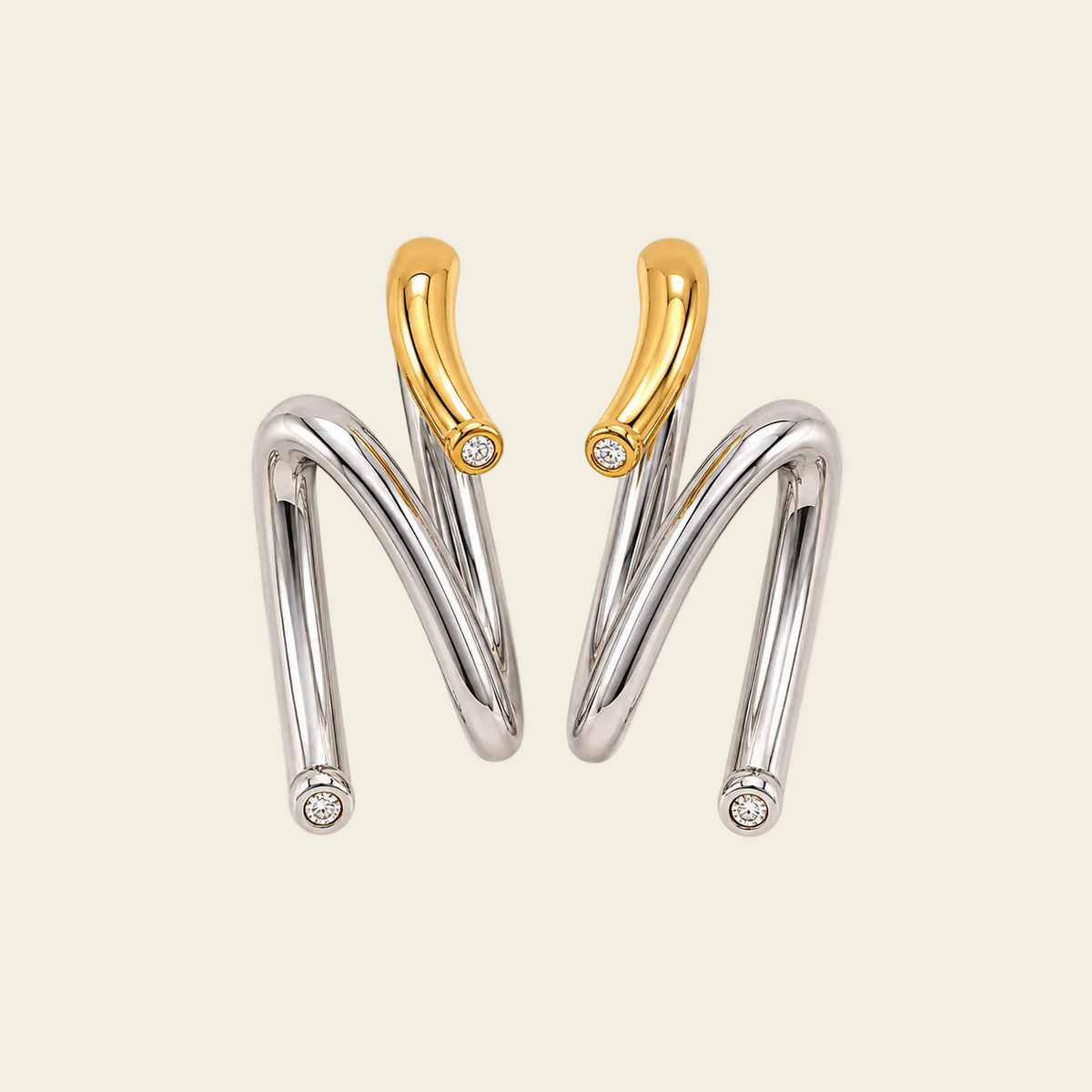 A pair of modern, abstract curved earrings in polished silver and yellow gold, each featuring a small round-cut clear gemstone at the tip and on the upper curve, set against a solid pale beige background.