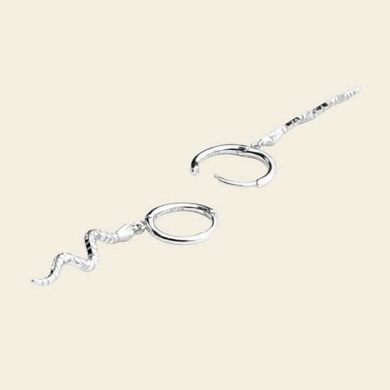 A pair of silver-toned ear cuffs with textured wavy dangling chains, displayed on a solid light beige background.