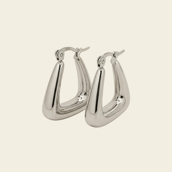A pair of polished silver-tone hoop earrings with a unique curved, teardrop-like shape and fishhook backings, displayed on a solid light beige background.