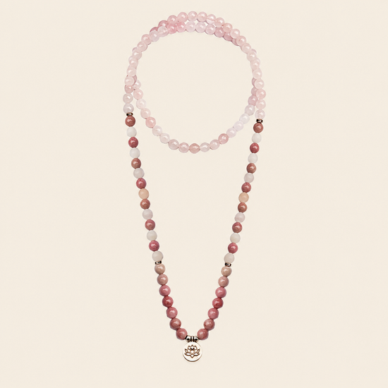 A double-strand beaded necklace featuring light pink and rose quartz beads, with small metallic spacer beads and a silver-toned lotus pendant at the bottom, displayed against a solid light beige background.