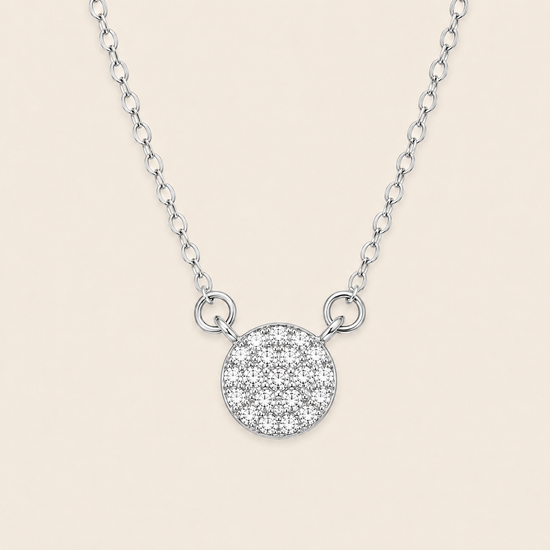 A silver chain necklace featuring a round pavé pendant with a textured, glittering surface, displayed against a solid light beige background.