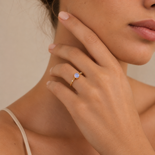 Close-up of a woman's hand wearing a gold ring with a blue gemstone, touching her face.