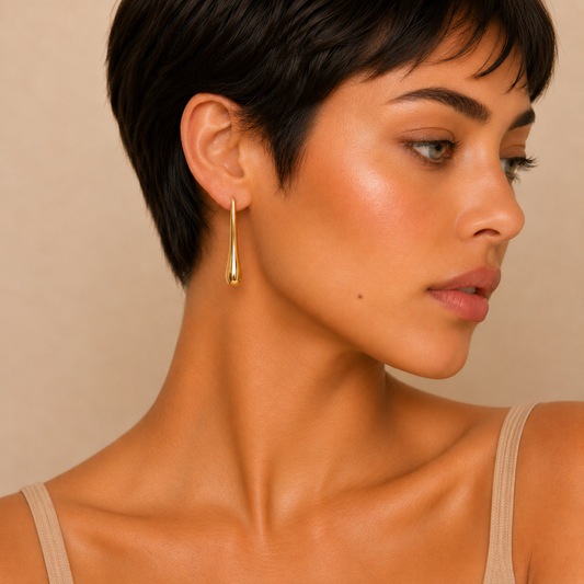 A close-up side profile of a woman with short dark hair wearing a delicate gold teardrop dangle earring against a warm beige background.