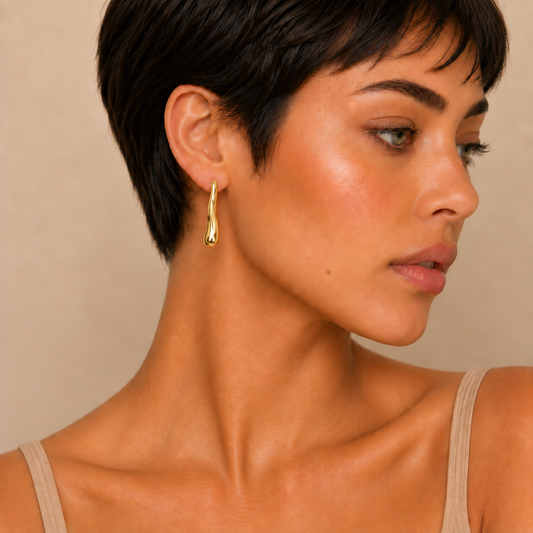 A close-up of a woman with short dark hair wearing a single gold teardrop-shaped dangle earring, shown in profile against a warm beige background.