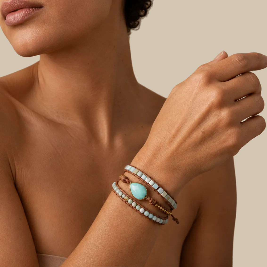 A close-up of a woman's wrist wearing a stack of bohemian-style bracelets, including a brown cord bracelet with a light blue teardrop stone and white beads.