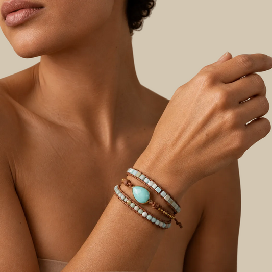 A close-up of a woman's wrist wearing a stack of bohemian-style bracelets, including a brown cord bracelet with a light blue teardrop stone and white beads.