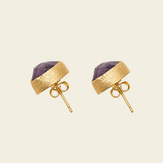 A pair of gold stud earrings featuring a textured gold band wrapping a faceted purple gemstone, shown on a light beige background.
