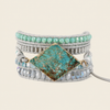 A multi-strand wrap bracelet featuring a large diamond-shaped turquoise stone centerpiece with gold-toned borders, surrounded by silver-toned beaded bands, light gray cord wraps, and rows of light blue faceted beads on a light beige background.