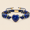 A beaded bracelet featuring a central heart-shaped lapis lazuli stone in a gold bezel, surrounded by blue and gold marbled beads, a navy blue cord, and gold-toned metal accents, set against a solid beige background.