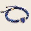 A beaded bracelet featuring deep blue and white marbled beads, a dark blue cord adjustable slider with gold-tone metal accents, and a polished heart-shaped blue stone charm set in gold-tone metal, displayed on a light beige background.
