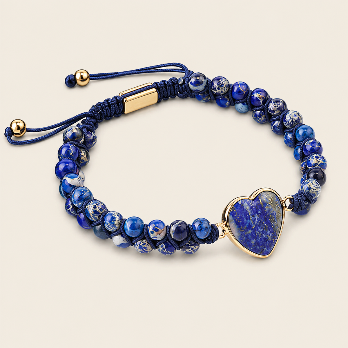 A beaded bracelet featuring deep blue and white marbled beads, a dark blue cord adjustable slider with gold-tone metal accents, and a polished heart-shaped blue stone charm set in gold-tone metal, displayed on a light beige background.