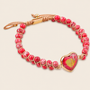 A beaded bracelet featuring textured red beads wrapped in gold-toned cord, with a gold heart-shaped charm containing a red and yellow marbled pattern, set against a light beige background.