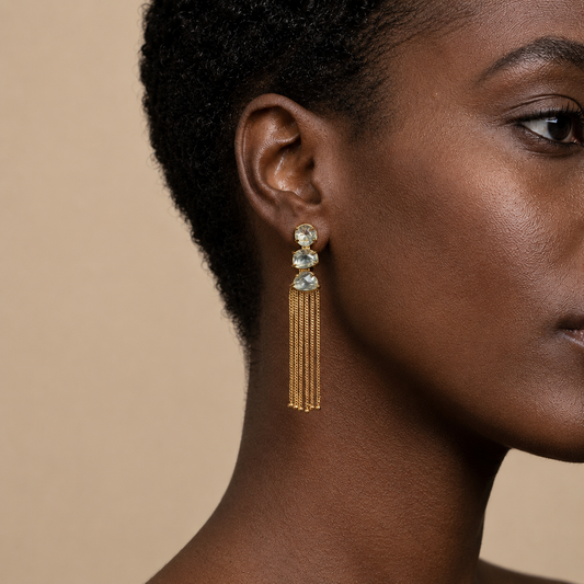 Close-up of a woman wearing gold tassel earrings with green gemstones on a beige background