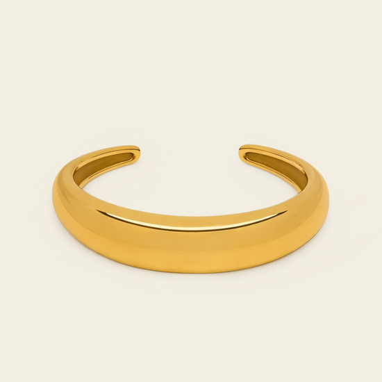 A polished gold open cuff bracelet, also known as a hinged bangle, shown on a solid light beige background.