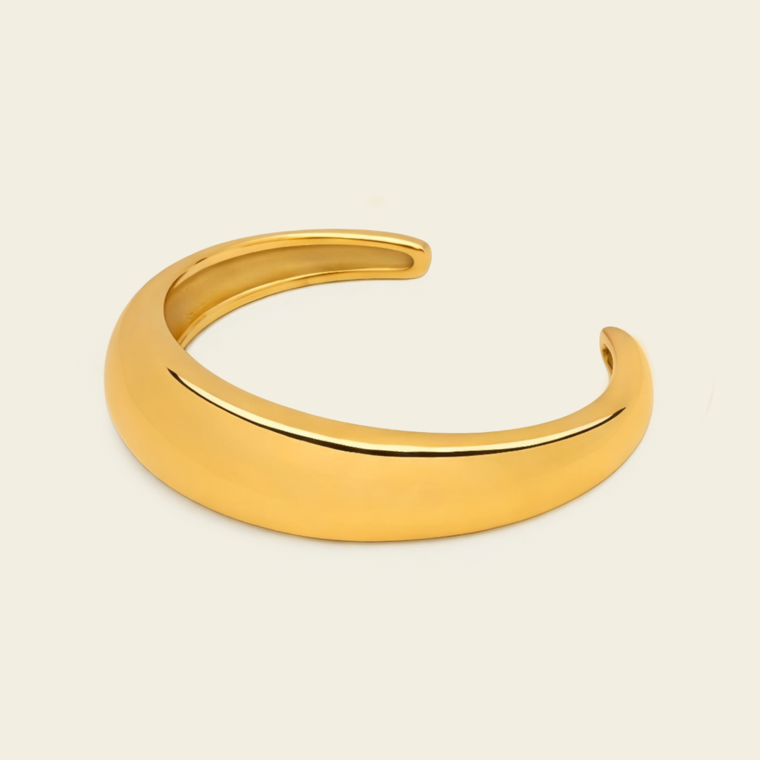 A polished gold open cuff bracelet, also known as a hinged bangle, shown on a solid light beige background.