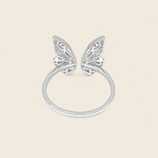 A silver butterfly ring with two delicate wing motifs on a smooth band, set against a solid light beige background.