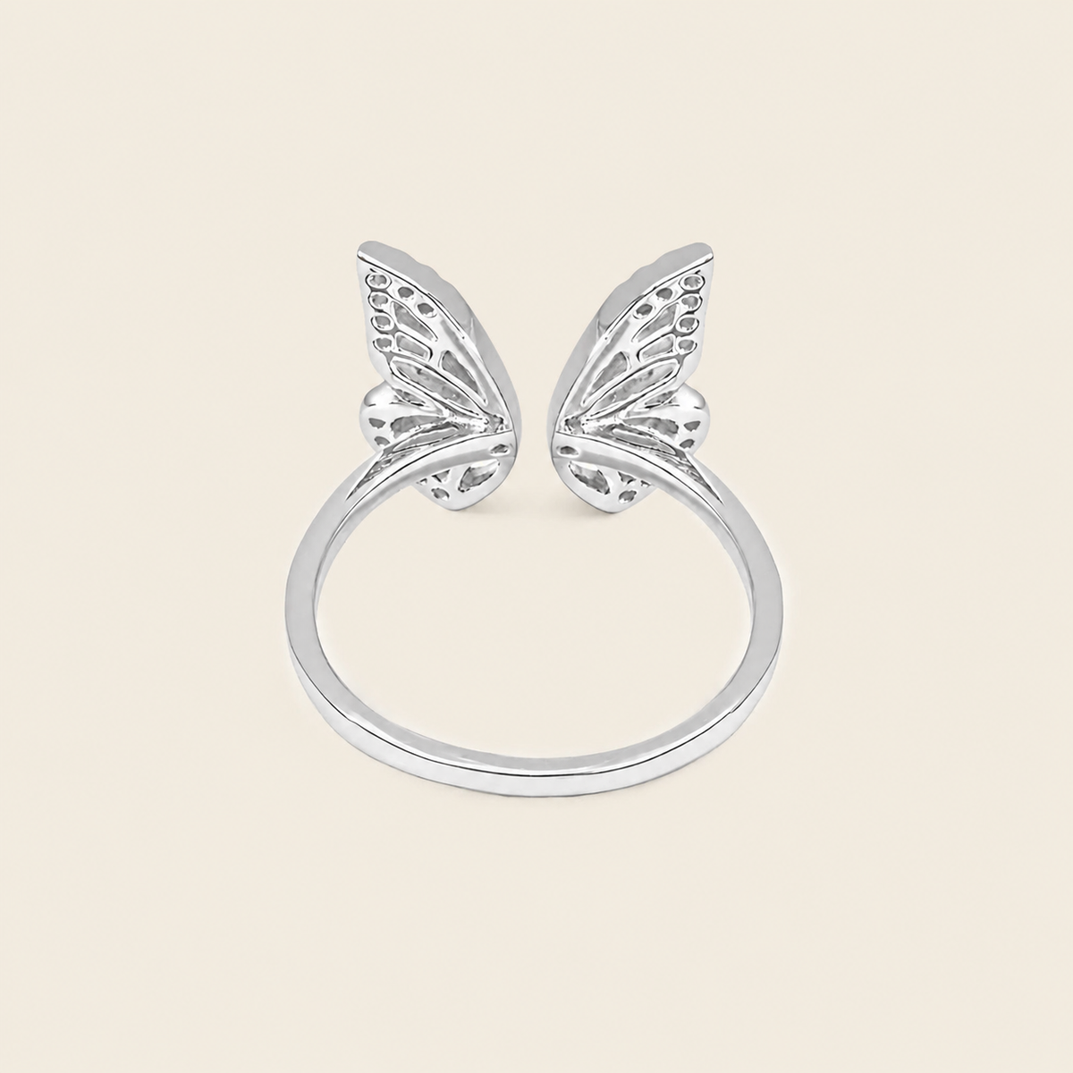 A silver butterfly ring with two delicate wing motifs on a smooth band, set against a solid light beige background.