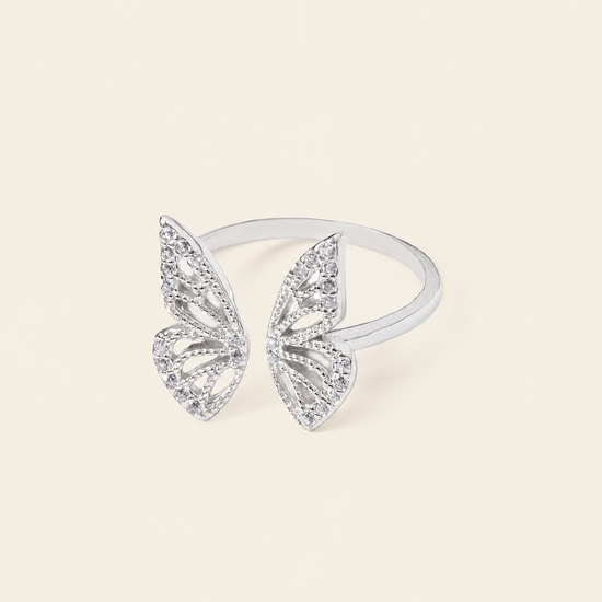 A silver butterfly ring set with clear pavé stones, displayed on a clean, solid cream-colored background.