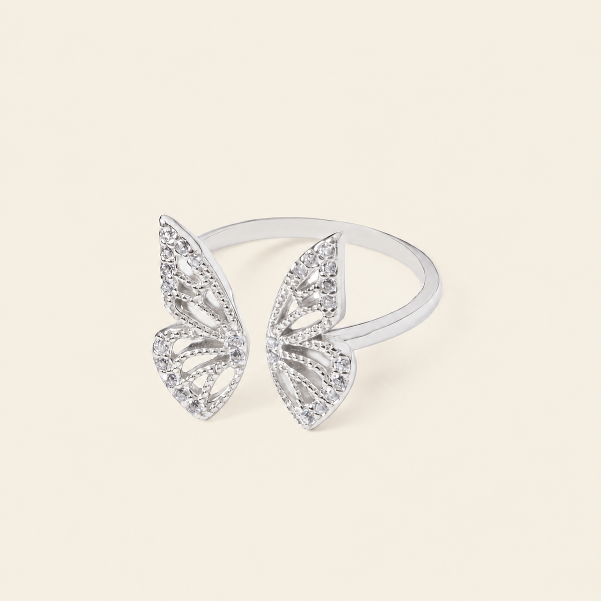 A silver butterfly ring set with clear pavé stones, displayed on a clean, solid cream-colored background.