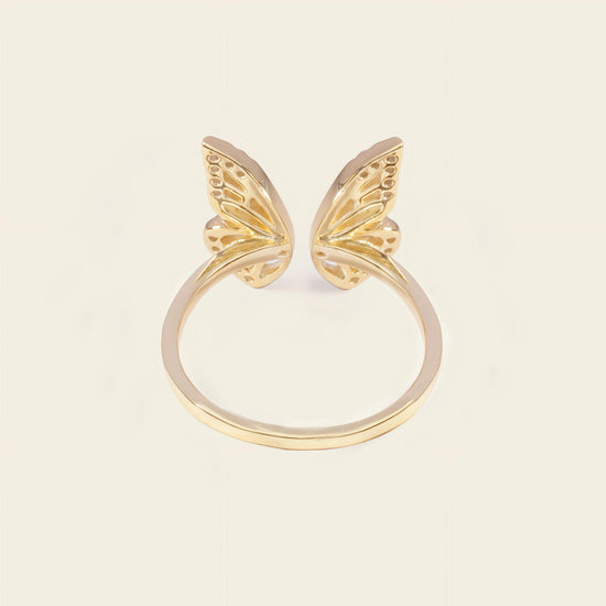 A gold plated butterfly ring with two delicate wing motifs on a smooth band, set against a solid light beige background.
