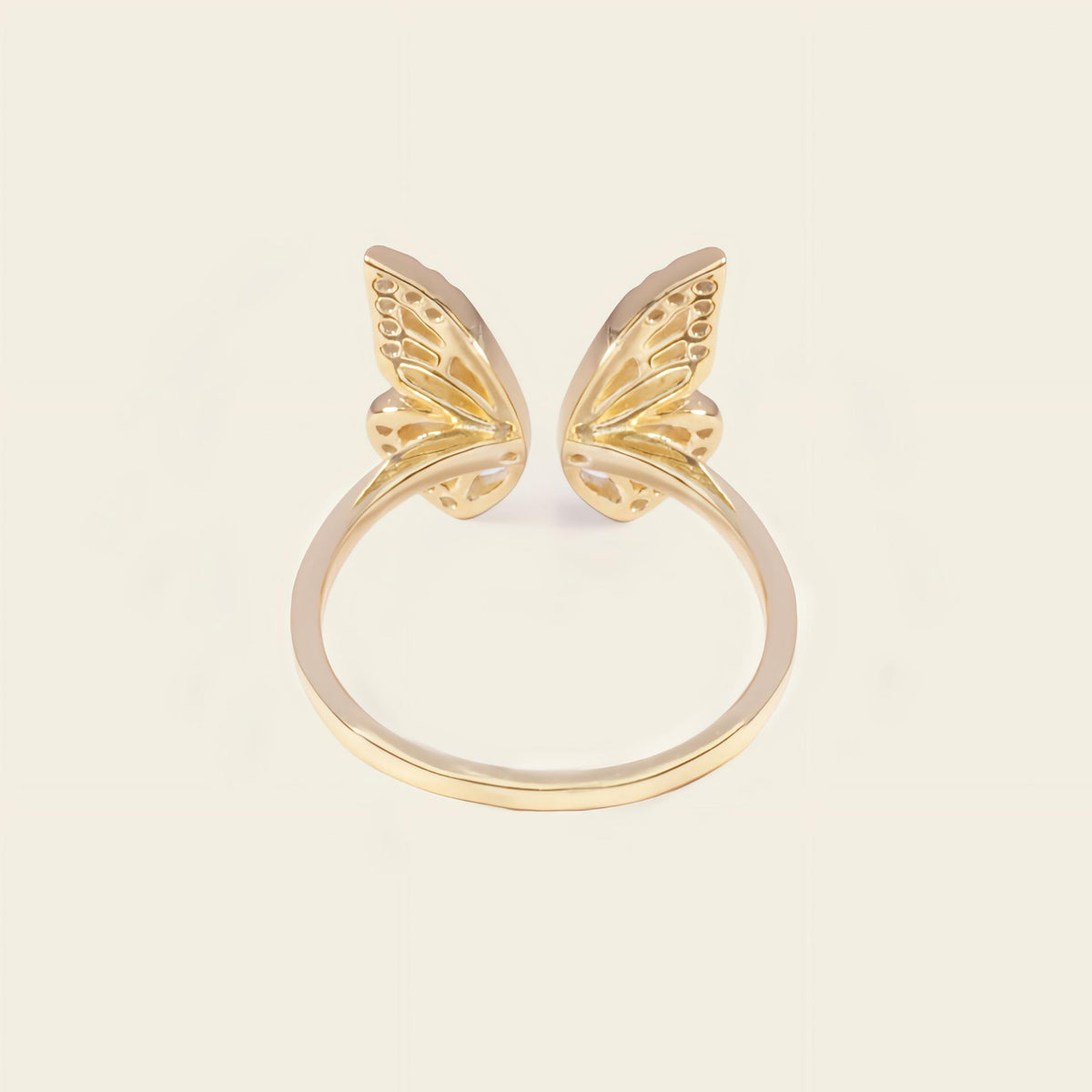 A gold plated butterfly ring with two delicate wing motifs on a smooth band, set against a solid light beige background.
