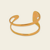A polished gold open cuff bracelet with a modern, flowing organic design, shown on a solid light cream background.