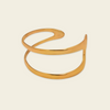 A polished gold-tone metal cuff bracelet with an organic, flowing looped design, shown on a solid light beige background.