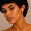 A close-up portrait of a woman with short curly dark hair wearing a small gold hoop earring. The background is a solid warm beige studio backdrop.