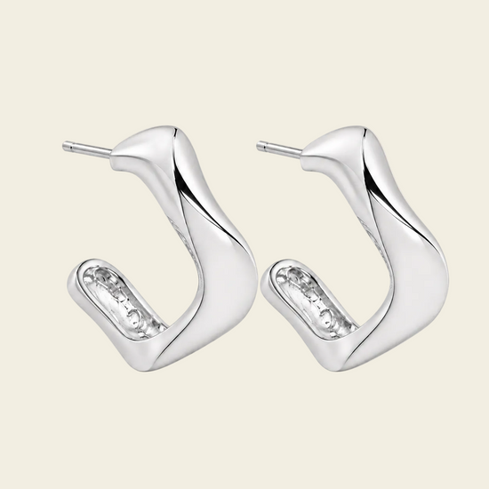 A pair of modern, polished silver huggie hoop earrings with a wavy, organic curve design and a small clear crystal accent on the inner band, set against a solid pale beige background.
