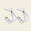 A pair of modern, polished silver huggie hoop earrings with a wavy, organic curve design and a small clear crystal accent on the inner band, set against a solid pale beige background.