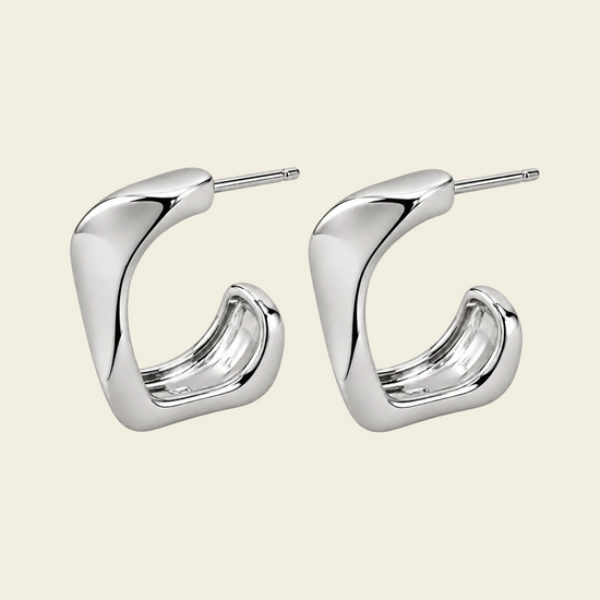 A pair of polished silver-tone geometric hoop earrings with a curved, sculptural design and straight post backs, displayed on a solid light beige background.