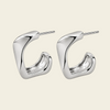 A pair of polished silver-tone geometric hoop earrings with a curved, sculptural design and straight post backs, displayed on a solid light beige background.