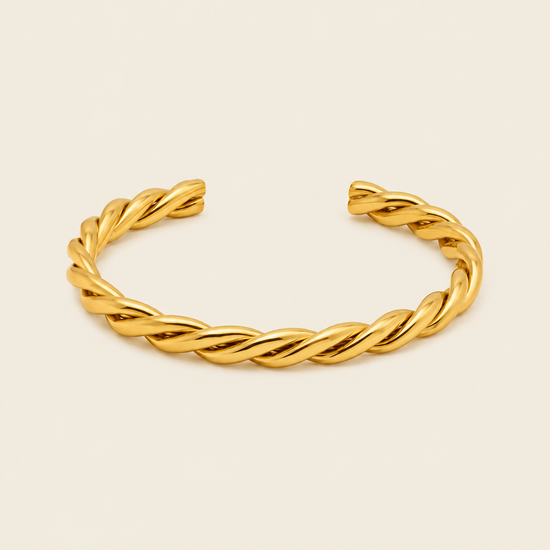 A twisted open cuff bracelet in polished yellow gold, displayed on a clean, solid, light cream background.