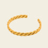 A twisted open cuff bracelet in polished yellow gold, displayed on a solid light beige background.