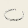 A twisted open cuff bracelet in polished silver, displayed on a solid light beige background.