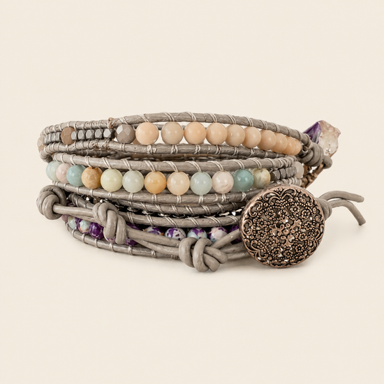 A stack of bohemian-style wrap bracelets featuring gray cord, natural stone beads in beige, green, and orange, purple crystal accents, and a large textured silver-toned metal disc clasp, set against a solid cream background.