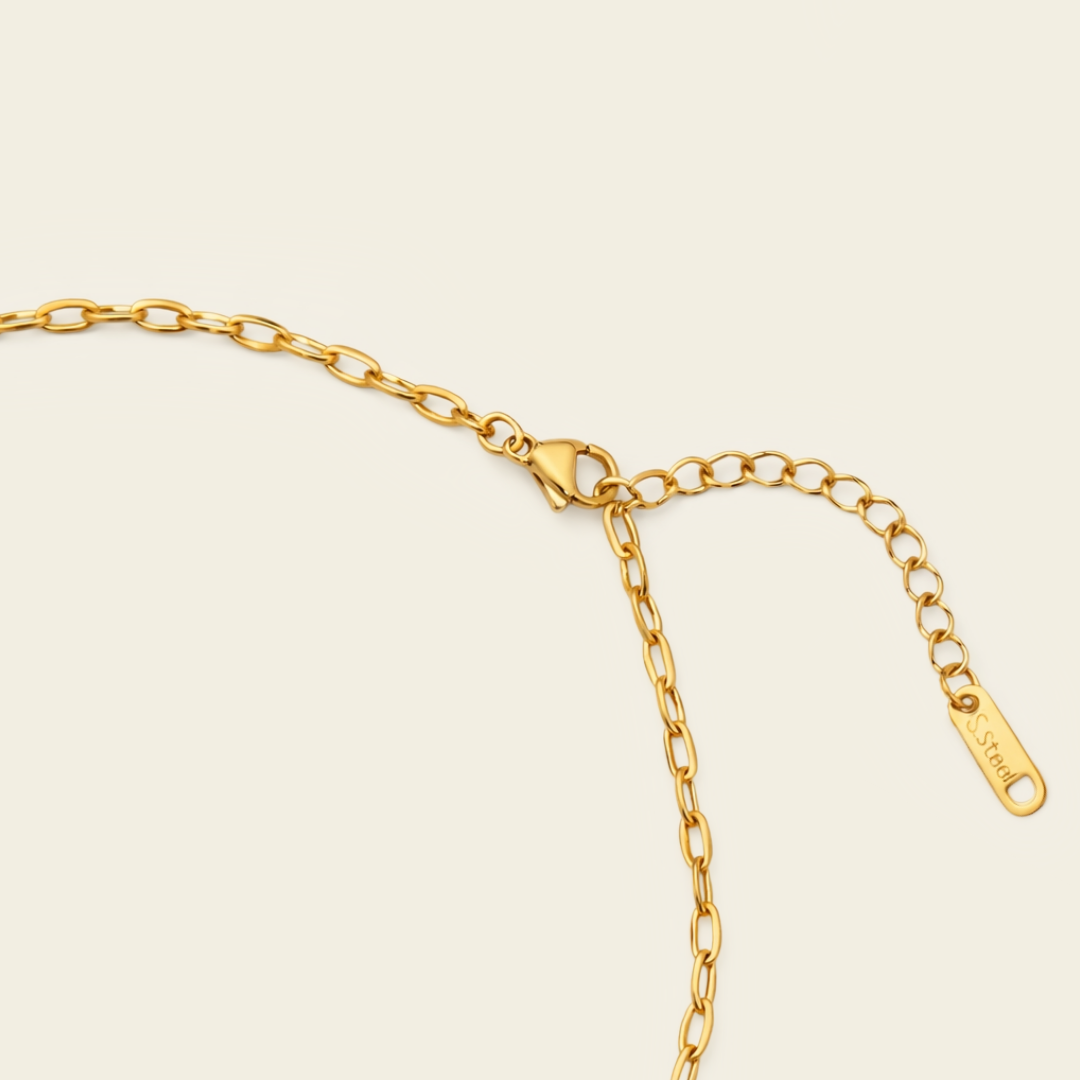 Close-up of a gold chain necklace with a lobster clasp, extender chain, and a small engraved tag on a light cream background.