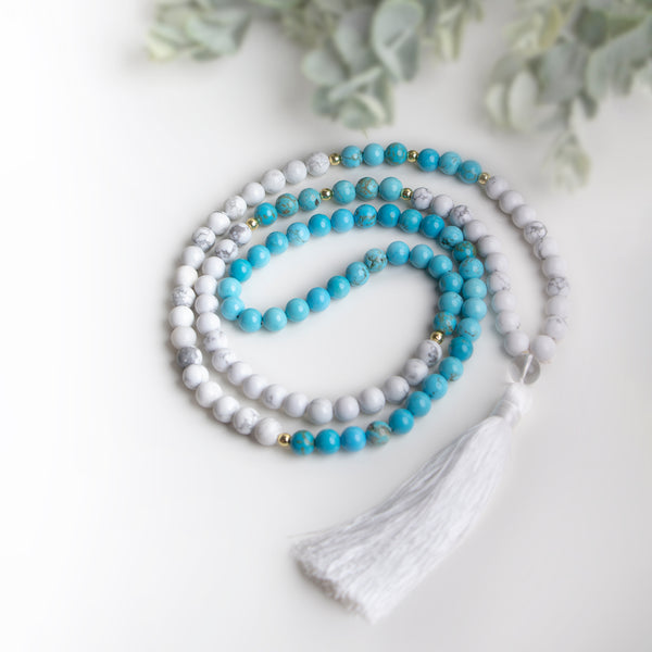  handmade necklace with white Howlite and turquoise beads, gold accents, and a soft white tassel, styled with greenery for a fresh boho look.