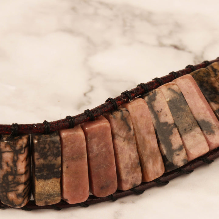 Close-up view of a handmade adjustable bracelet featuring natural Rhodonite tube stones in shades of pink and black, secured with dark leather cord.